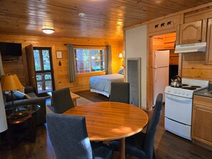 A cabin interior with a bed, dining table, chairs, a kitchen with a stove, and a TV.