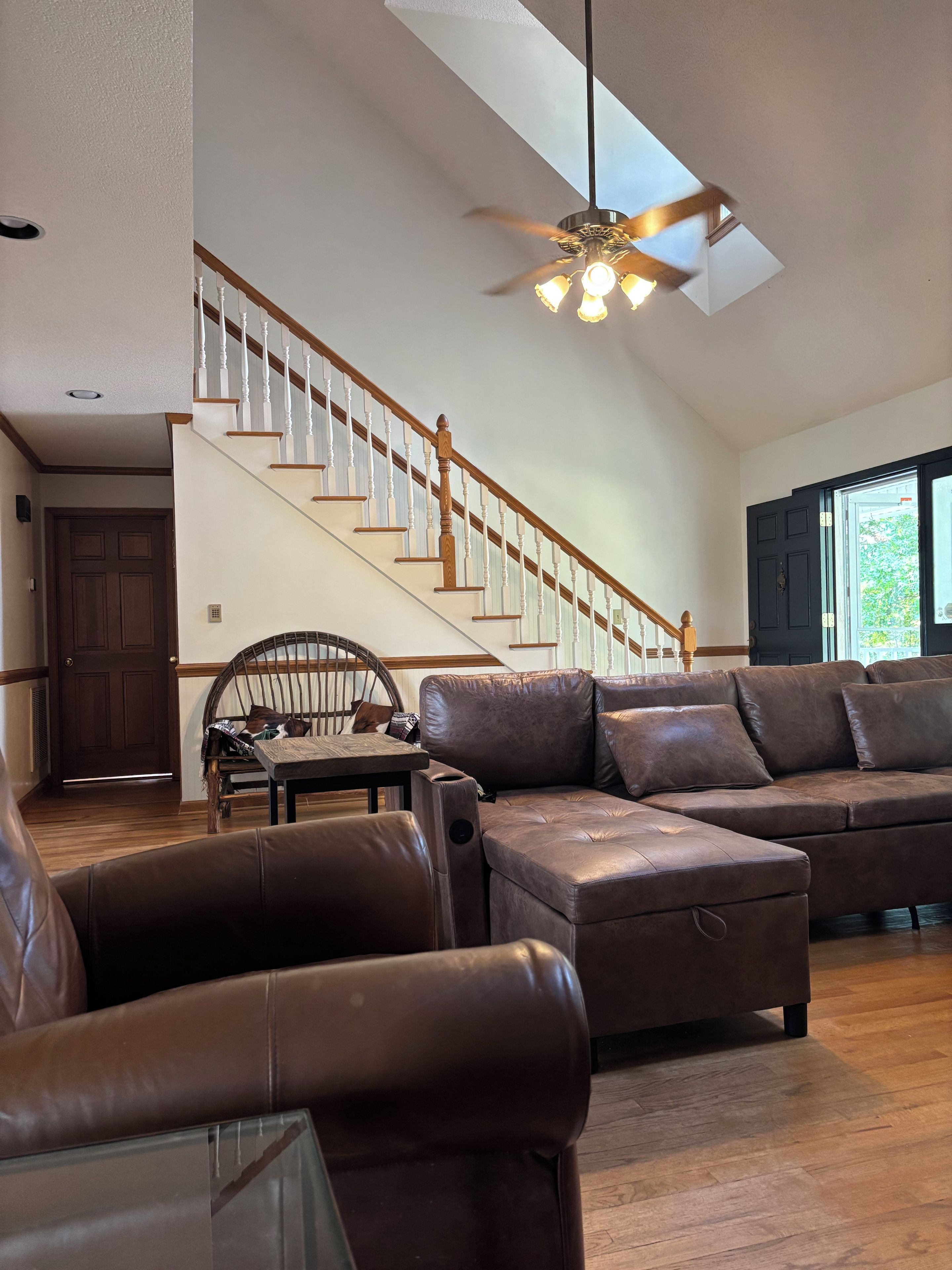 A spacious living room with a high ceiling, a wooden staircase, leather sofas, and a ceiling fan.