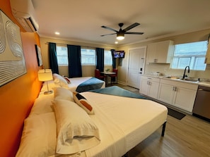 Quarto de hotel com duas camas, cozinha compacta, ventilador de teto e televisão na parede.
