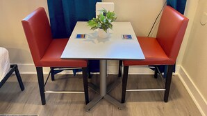 Una habitación con una mesa blanca, dos sillas rojas, una planta en maceta y una ventana con persianas.