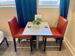 Quarto com uma mesa branca, duas cadeiras vermelhas, um vaso com planta e uma janela com persianas.