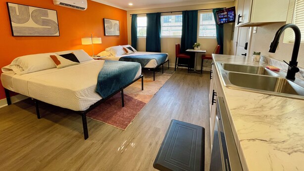 Una habitación de hotel compacta con dos camas, una kitchenette, un ventilador de techo y un televisor.