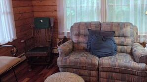 A cozy cabin interior with a wooden ceiling, a patterned sofa, a round ottoman, and a wooden chair. - Cozy lakefront cabin retreat in Greensboro, Vermont perfect for your getaway (Greensboro)
