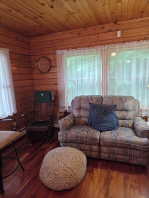 A cozy cabin interior with a wooden ceiling, a patterned sofa, a round ottoman, and a wooden chair. - Cozy lakefront cabin retreat in Greensboro, Vermont perfect for your getaway (Greensboro)