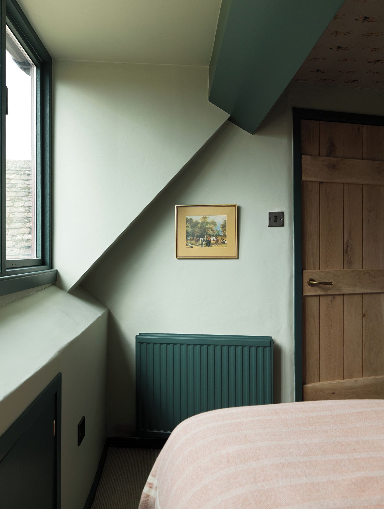 Una habitación con techo inclinado, un radiador, un cuadro enmarcado en la pared, una puerta y una ventana.