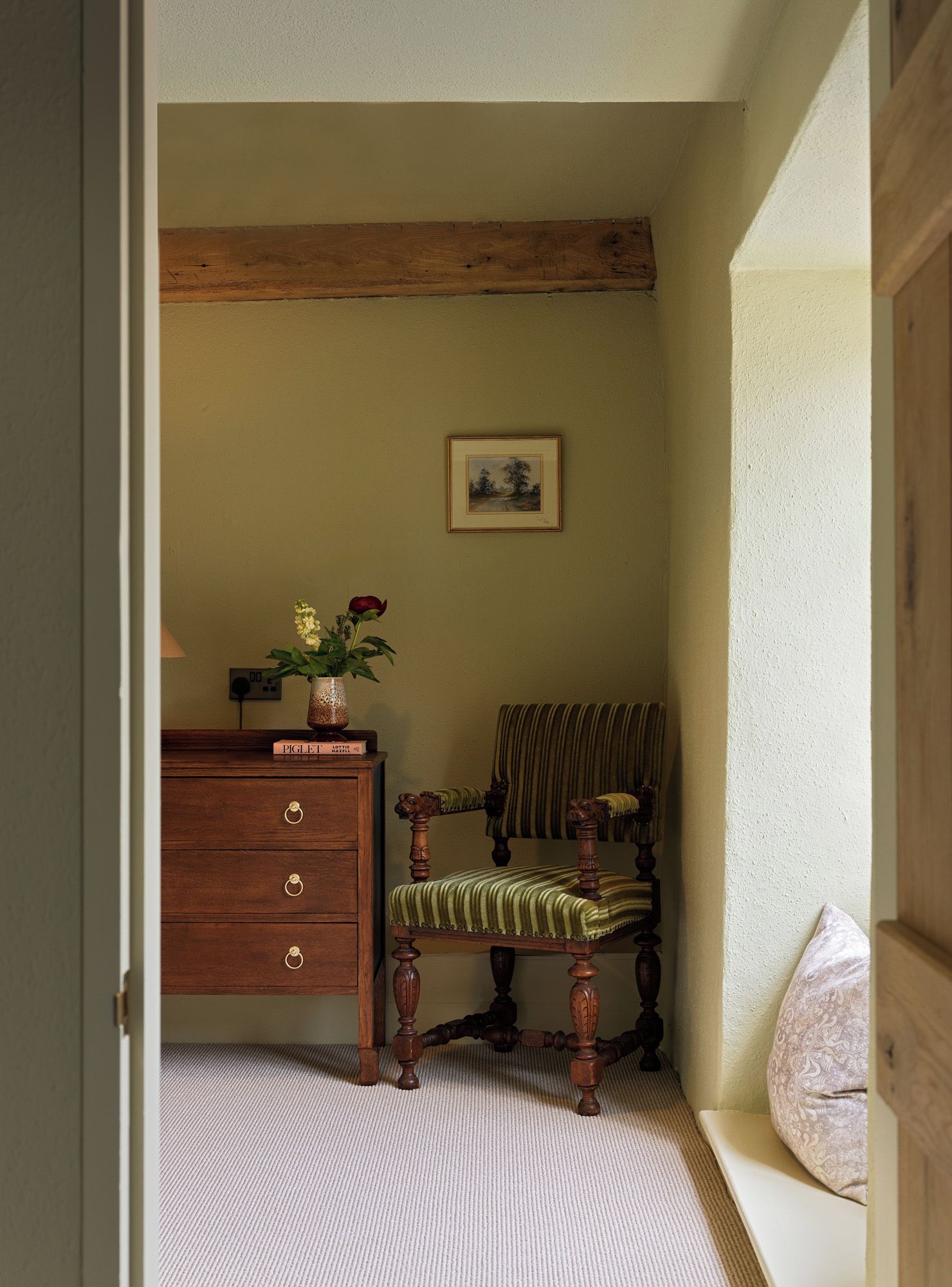 A corner room with a wooden dresser, a chair with a patterned cushion, a framed picture on the wall, and a vase with flowers on the dresser.
