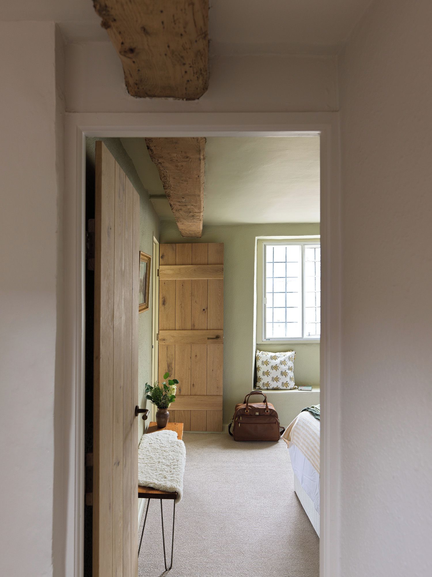 A room with a wooden door, a bench, a window, a bed, and a bag.