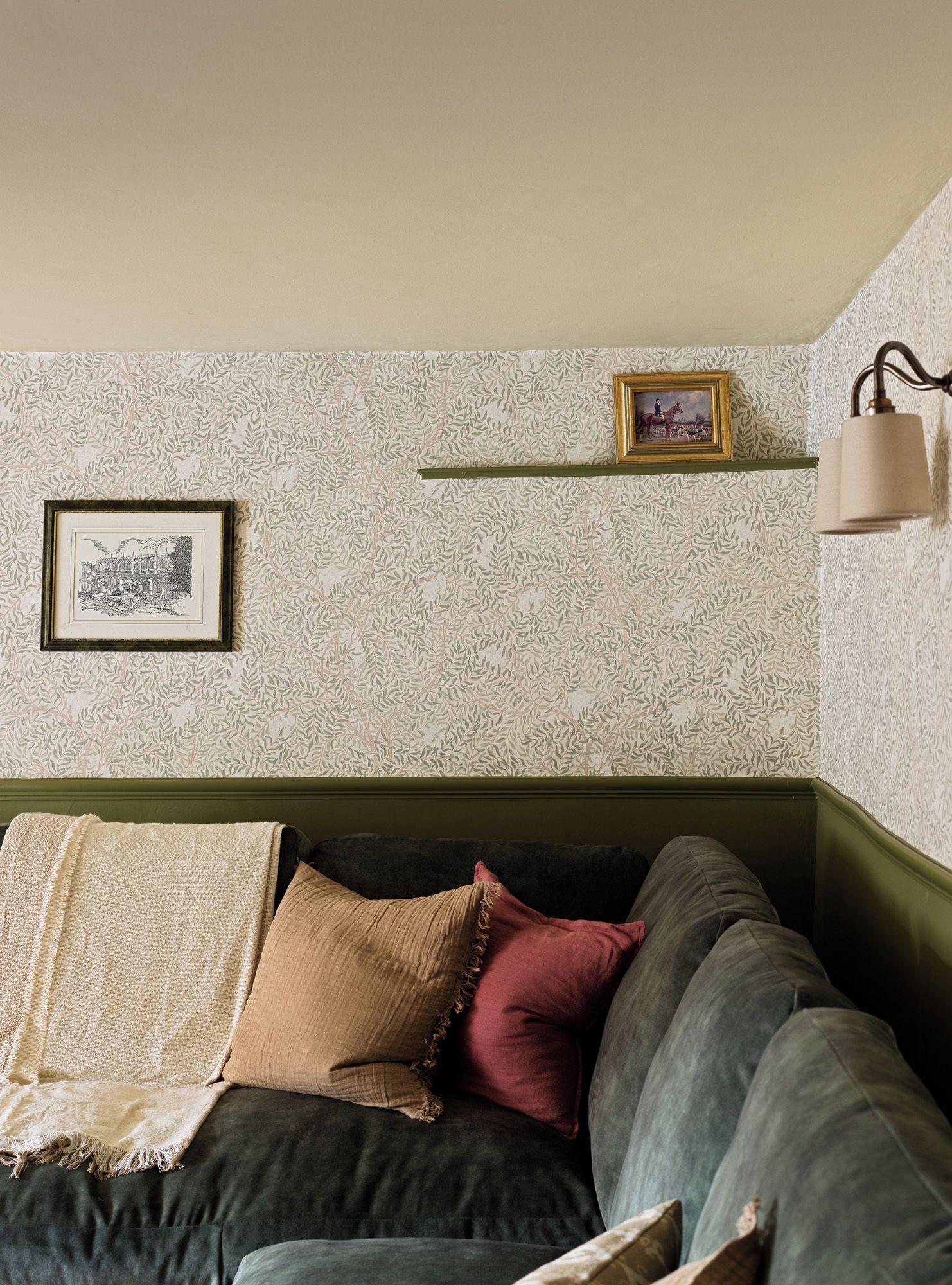 Een kamer met een groen stoffen bank, behang met een patroon, een ingelijst schilderij aan de muur en een wandlamp.