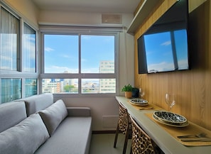 A modern living room with a sofa, dining table, and chairs, a flat-screen TV mounted on the wall, and large windows offering a city view. - Fully furnished apartment 180m from the beach. (Salvador)