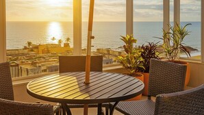 Ein Balkon mit rundem Tisch und Stühlen, der auf einen Küsten-Stadtbild im Sonnenuntergang blickt.