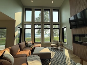 A spacious living room with a large window overlooking a lake, a comfortable sofa, a small table, and a flat-screen TV mounted on the wall. - Modern 4 bedroom Lakehouse on Alexandria chain of lakes! (Alexandria)