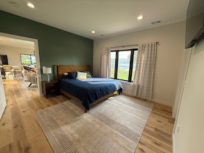 A bedroom with a bed, a nightstand, a window with curtains, a view of greenery outside, and a dining area in the background. - Modern 4 bedroom Lakehouse on Alexandria chain of lakes! (Alexandria)