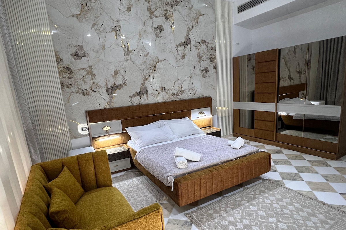 Une chambre à coucher moderne avec un grand lit, une table de chevet, une chaise, un miroir et un tapis à motifs.