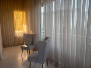 Una habitación con dos sillones grises, un ventanal con cortinas traslúcidas y suelo de madera.
