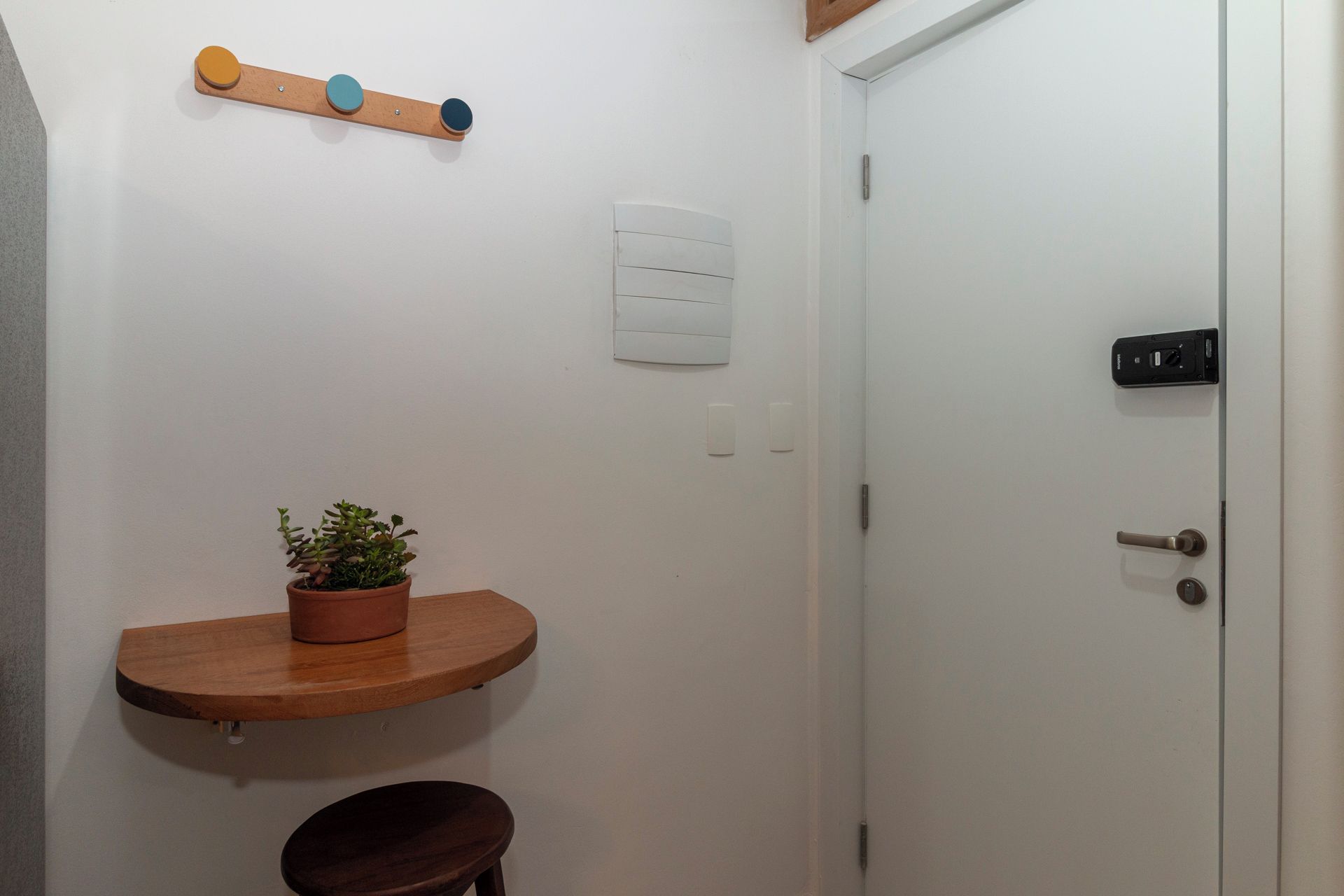 Un baño pequeño con un estante de madera que sostiene una planta en maceta, una puerta blanca con pomo negro y un interruptor de luz en la pared.