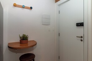 Un baño pequeño con un estante de madera que sostiene una planta en maceta, una puerta blanca con pomo negro y un interruptor de luz en la pared.