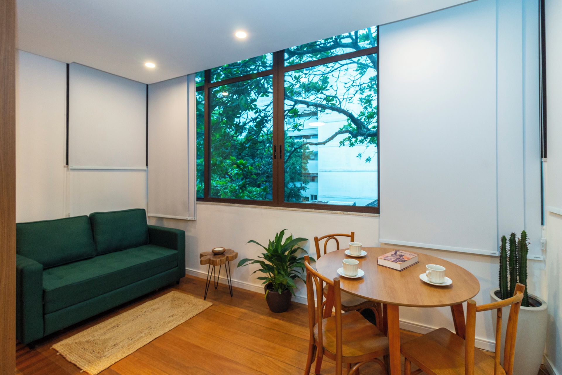 Een kamer met een groene bank, een houten eettafel met stoelen, een potplant en een groot raam met uitzicht op bomen en gebouwen.