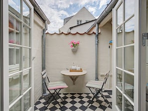 Une petite terrasse avec un sol à carreaux, deux chaises, une table avec une théière et un pot de fleurs accroché au mur.