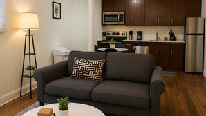 A modern living room with a dark grey sofa, a white coffee table, and a floor lamp. A kitchen with dark wooden cabinets, stainless steel appliances, and a white countertop is visible in the background. - Delightful 1BR Apt in Ravenswood (Chicago)