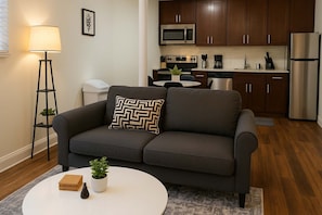 A modern living room with a dark grey sofa, a white coffee table, and a floor lamp. A kitchen with dark wooden cabinets, stainless steel appliances, and a white countertop is visible in the background. - Delightful 1BR Apt in Ravenswood (Chicago)