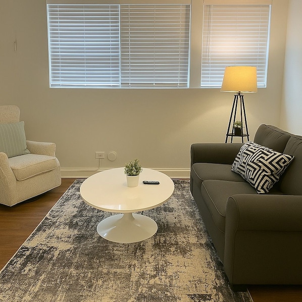 A living room with a round coffee table, a sofa, a chair, a lamp, and a window with blinds. - Delightful 1BR Apt in Ravenswood (Chicago)