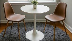 Une pièce avec une table ronde blanche, deux chaises marron, un tapis gris à poils longs et une plante en pot posée sur la table.