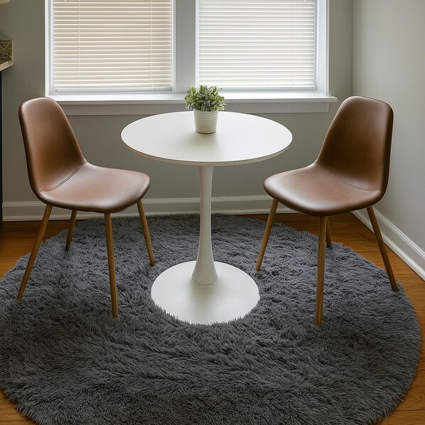A room with a round white table, two brown chairs, a grey furry rug, and a potted plant on the table.