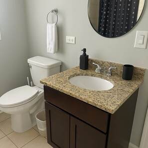 Un baño con inodoro blanco, lavamanos con encimera de granito, dispensador de jabón negro, taza negra, toalla blanca y espejo redondo.