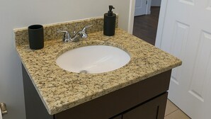 Une salle de bain avec un lavabo doté d’un plan de travail en granit, un miroir, un robinet, un distributeur de savon et un porte-rouleau de papier toilette.