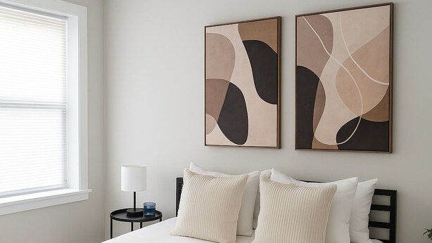 Une chambre avec un grand lit, deux tables de chevet, une lampe, des œuvres d’art abstraites accrochées au mur et une fenêtre avec des stores.