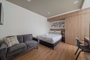 Ein modernes Schlafzimmer mit einem grauen Sofa, einem Bett mit weißer Bettwäsche, einem Holzschrank und einem Schreibtisch mit Stuhl.