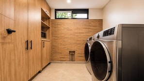 Une buanderie avec des armoires en bois, un mur en briques, ainsi qu’un lave-linge et un sèche-linge en acier inoxydable.