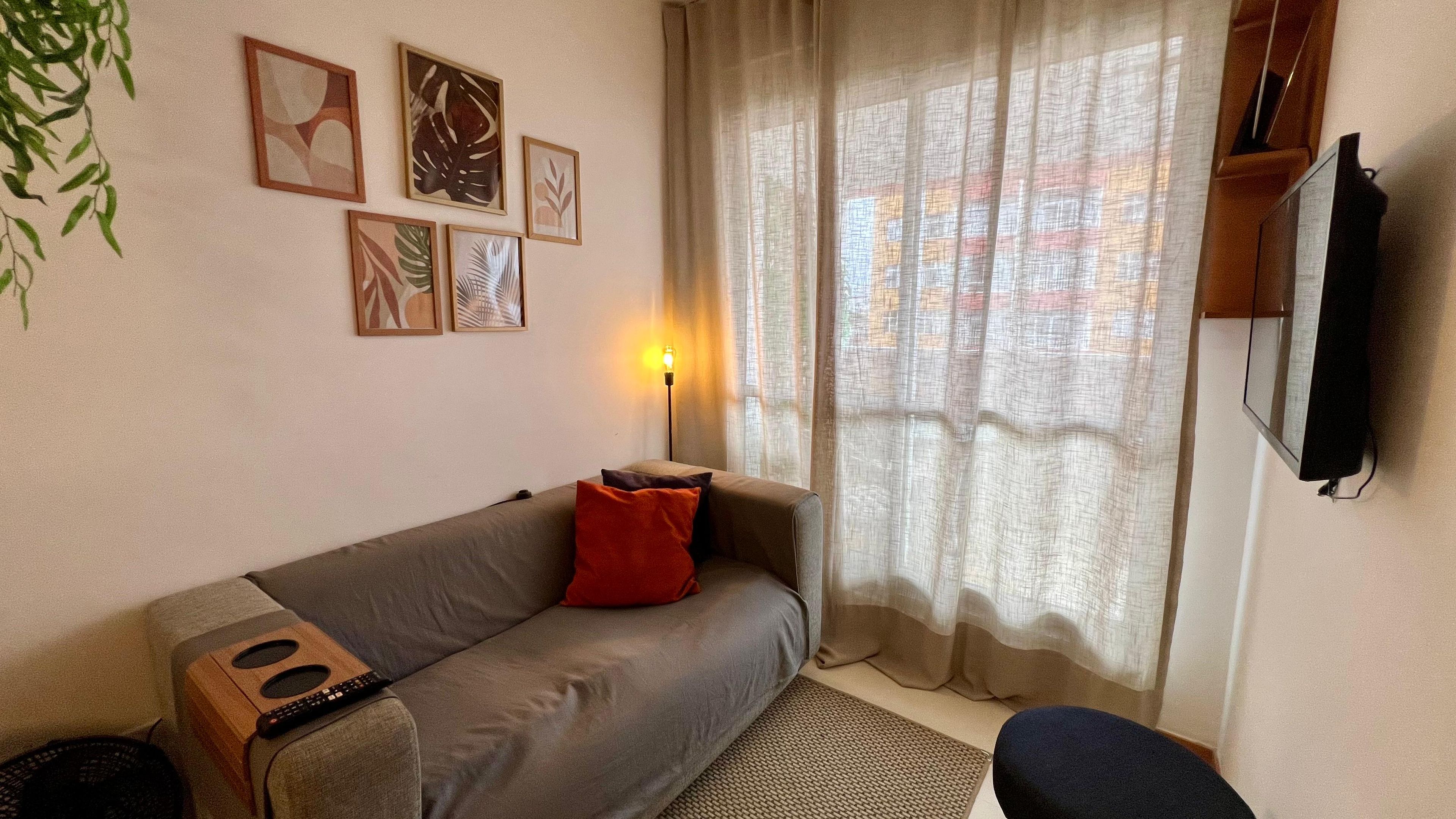 A living room with a grey sofa, a red pillow, a wall-mounted TV, and framed artwork.