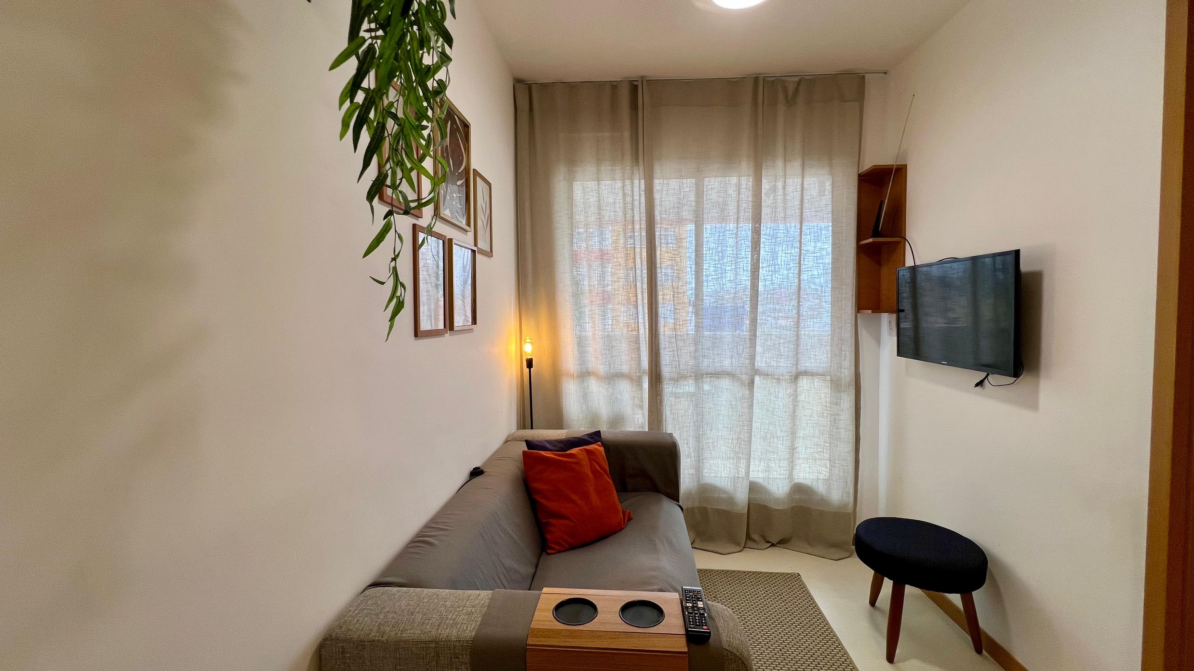 Uma sala de estar com sofá, uma mesinha, televisão e janela com cortinas.