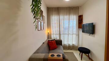 Uma sala de estar com sofá, uma mesinha, televisão e janela com cortinas.
