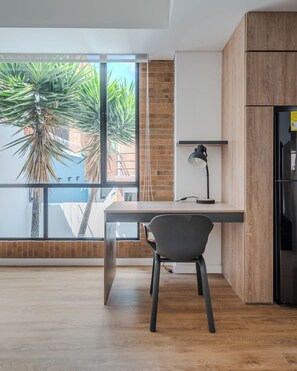 Une cuisine moderne avec des meubles en bois, un réfrigérateur noir, un bureau avec une chaise et une grande fenêtre donnant sur des palmiers.