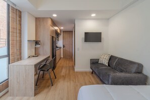 Habitación de hotel compacta con kitchenette, sofá, escritorio con silla y televisor montado en la pared.