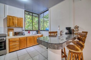 A kitchen with wooden cabinets, a microwave, a coffee maker, and a dining area with a glass table and wooden chairs. - Haze Villa | Tropical Retreat & Pool (Puerto Viejo De Talamanca)