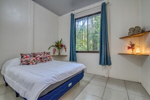 Un dormitorio con cama, ventana con cortinas, estante junto a la cama con toallas y una planta en maceta.