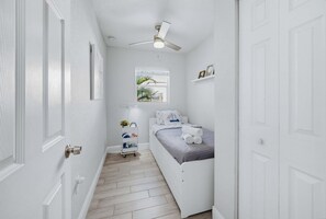Ein schmaler Flur führt zu einem Schlafzimmer mit Bett, Deckenventilator, Fenster mit Aussicht und einem kleinen Regal mit Gegenständen.