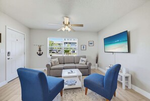 Ein Wohnzimmer mit einer Couch, zwei Sesseln, einem Couchtisch, einem Deckenventilator, einem Fernseher und einem Fenster mit Blick auf ein Gebäude und Bäume.
