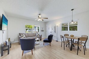 Ein geräumiges Wohnzimmer mit einer Couch, Sesseln, einem Couchtisch, einem Esstisch mit Stühlen, einem Deckenventilator und einem an der Wand befestigten Fernseher.