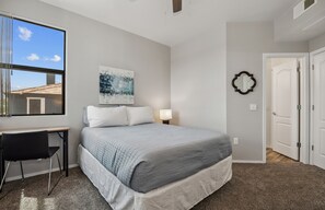 Ein Schlafzimmer mit einem Bett, einem Schreibtisch und einem Stuhl. Es gibt ein Fenster, ein Bild an der Wand und eine Tür.