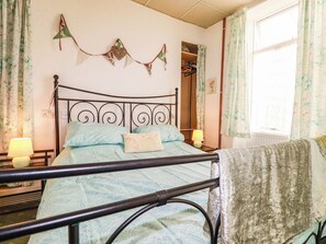 Ein Schlafzimmer mit einem Metallbettgestell, einem Nachttisch mit Lampe, einem Fenster mit floralem Vorhang und einer Wanddekoration.