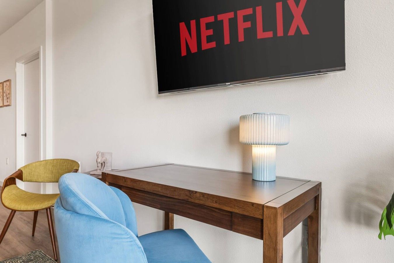 A room with a wooden desk, a lamp, a chair, and a wall-mounted TV displaying the Netflix logo.