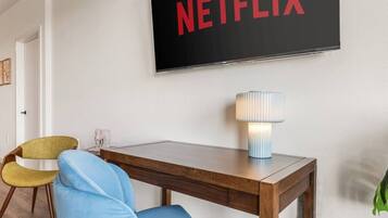 A room with a wooden desk, a lamp, a chair, and a wall-mounted TV displaying the Netflix logo.