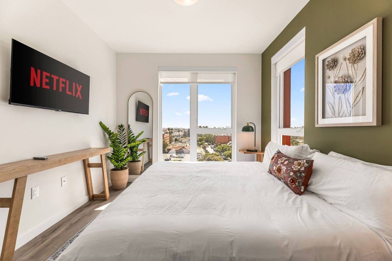 A bedroom with a large bed, a TV mounted on the wall, a desk with a plant, and a view of the city through a window.