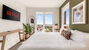 A bedroom with a large bed, a TV mounted on the wall, a desk with a plant, and a view of the city through a window.
