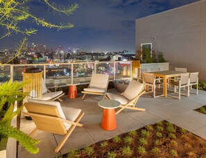 Terraço no telhado com áreas de estar, uma mesa de jantar e vista para a cidade à noite.
