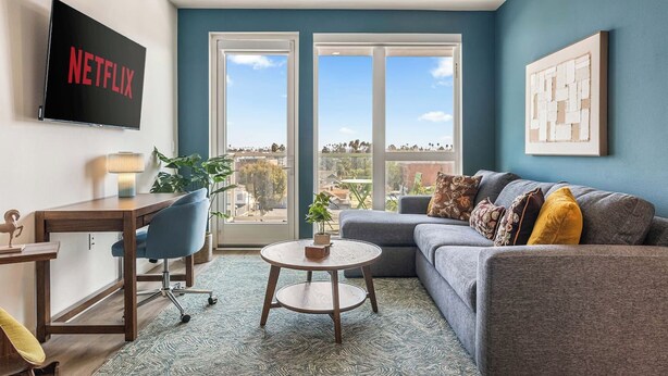 A modern living room with a grey sofa, a coffee table, a desk with a chair, a TV mounted on the wall, and a large window with a view of the city. - Light-Filled K-town 3BD 3BTH W/Pool, Gym & Parking (Los Angeles)